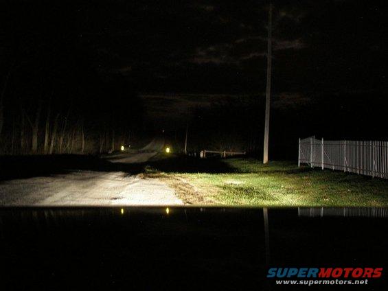View down a dark road at night, illuminated by headlights. Trees line the left, a fence on the right
