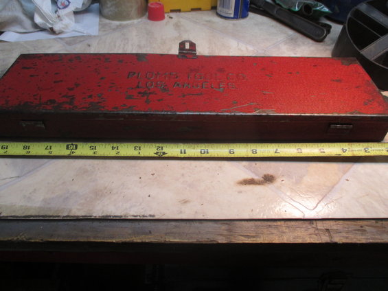 Red metal tool box with 'PLOMB TOOLS LOS ANGELES' embossed on lid. Tape measure below shows 11-19 inches