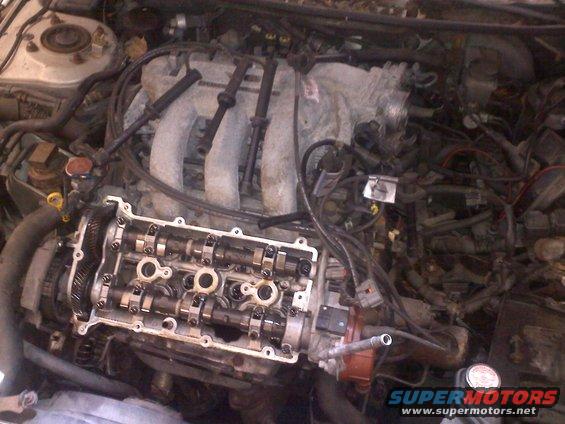 Engine bay with valve cover removed, showing camshafts and spark plug wires. Watermark reads "SUPERMOTORS.NET"