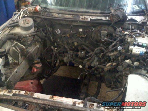 Car engine bay with engine and wiring harness exposed, appears to be undergoing maintenance or repair