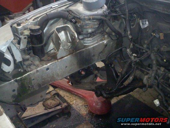 Car engine bay with exposed metal frame, hoses, and wiring. A red jack supports the chassis