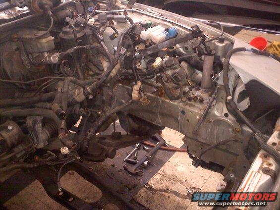 Disassembled car engine bay with exposed wiring and engine components. Tools are on the floor
