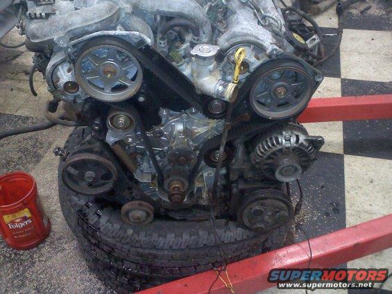 Engine assembly with belts and pulleys, resting on a tire. A red Folgers coffee can is nearby
