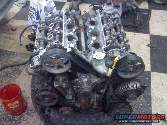 Engine block with exposed camshafts and timing belts, resting on a tire. Red Folgers coffee can nearby