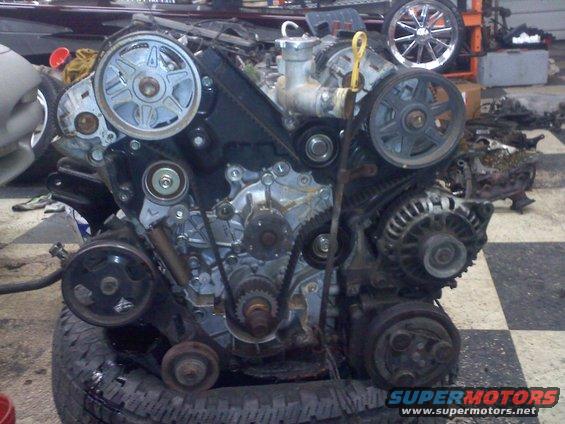 Engine assembly with belts, pulleys, and alternator. Water pump visible at top center. Supermotors.net logo at bottom right