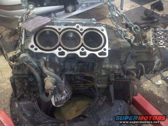 Engine block with three cylinders visible, suspended by chains. A tire is visible below
