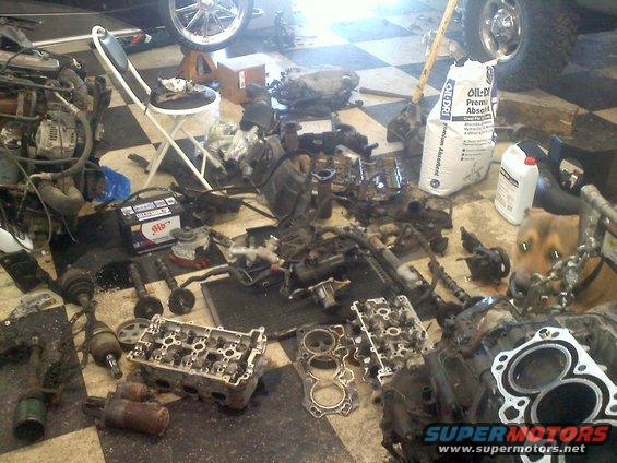 Disassembled engine parts and a car battery on a checkered floor, with a dog in the background