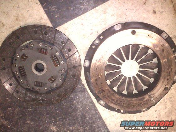Clutch disc and pressure plate on checkered floor. The disc has springs and a splined center. The pressure plate has radiating fins