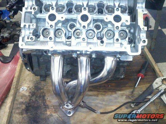 Engine block with chrome exhaust manifold and oxygen sensor attached, resting on a wooden surface with tools
