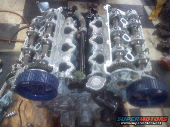 Engine block with exposed camshafts and blue timing gears. Some hoses and metal components are visible