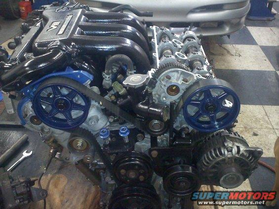 Engine with black intake manifold and blue pulleys, partially visible car front in background