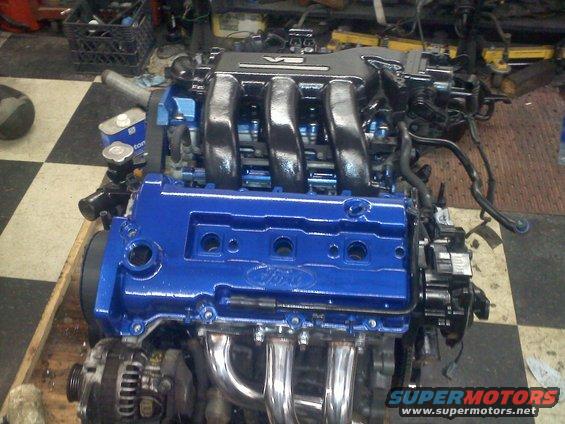 Ford V6 engine, blue painted valve covers, chrome exhaust manifold. Supermotors.net logo visible
