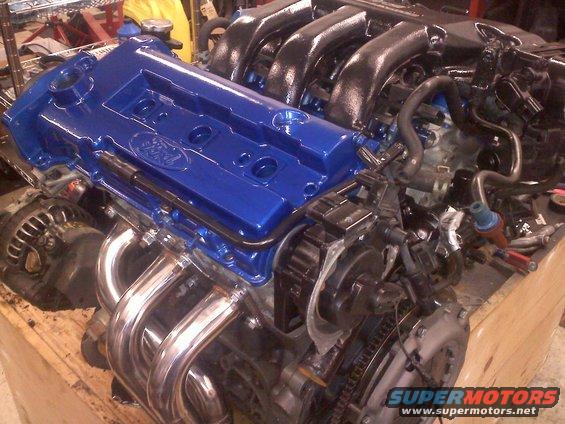 Blue Ford engine valve cover with Ford logo, chrome exhaust headers, and black intake manifold