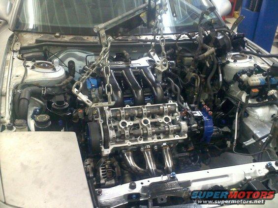 Engine being lifted out of a car with chains and an engine hoist. The engine has exposed camshafts and exhaust headers