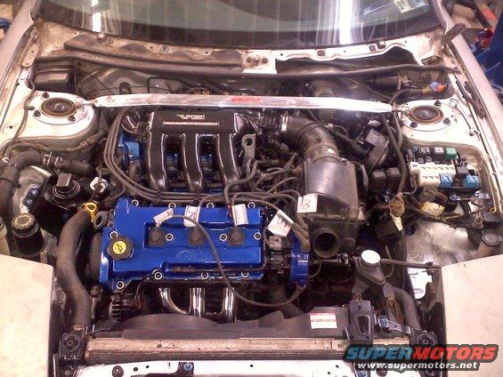 Engine bay with blue valve covers and chrome headers. A silver strut bar is visible above the engine