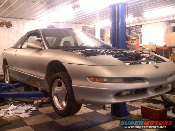 Silver 1993 Ford Probe GT on a car lift, hood open, showing engine bay. Garage setting