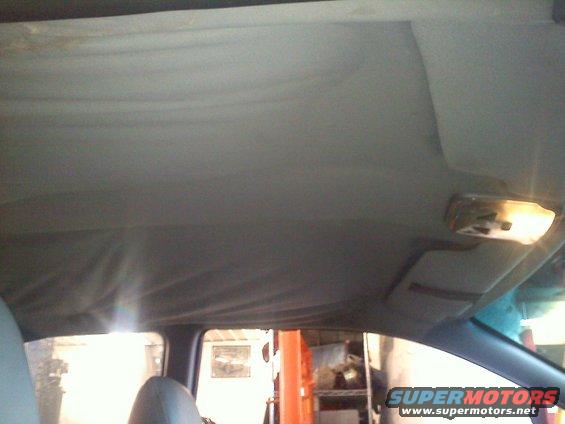 Car interior ceiling with sagging fabric and a dome light. A sun visor is visible