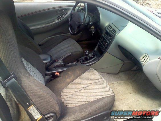Car interior with fabric seats, dashboard, steering wheel, and gear shift. Some dirt is visible on the seats and floor