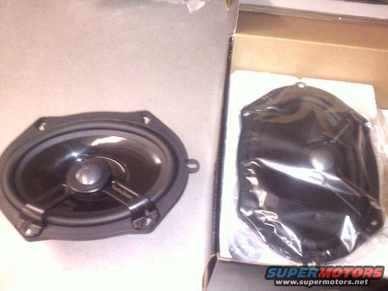 Oval car speaker next to a black plastic mounting bracket in a box