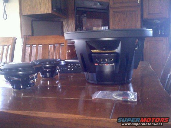 Car audio subwoofers and amplifier on a wooden table. One large subwoofer is in the foreground, with two smaller speakers and an amplifier behind it