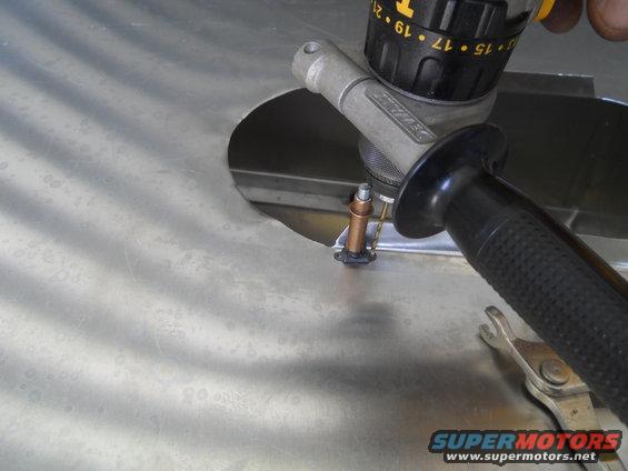 Drill bit entering a circular hole in a brushed metal panel, with a wrench visible below