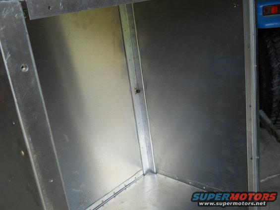 Inside of a metal cargo area with visible rivets and seams. Part of a blue vehicle and tire visible on the right