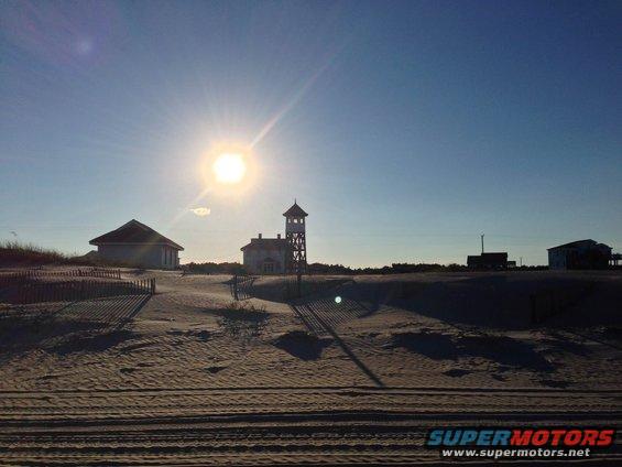 beach with sand dunes, a lighthouse-like tower, and buildings under a bright sun