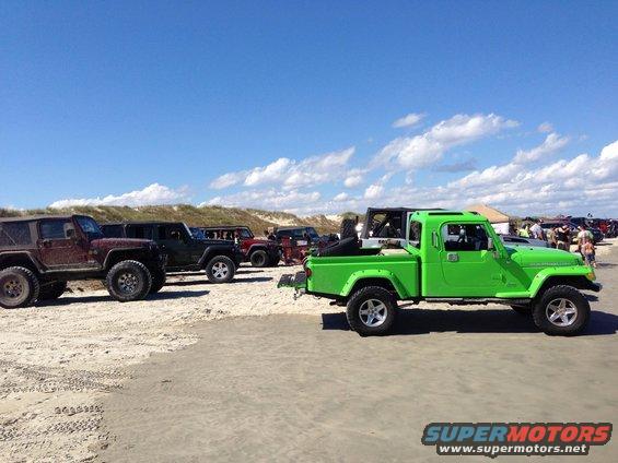 Green Jeep pickup truck parked on sand with other Jeeps in background