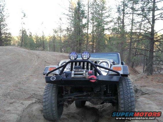 Front view of a silver Jeep Wrangler on a dirt trail with trees in the background. It has a winch and off-road lights