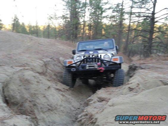 2005 Jeep Wrangler driving through a rutted dirt trail with trees in the background