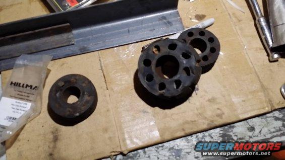 Three rubber isolators and metal brackets on a cardboard surface. A bag labeled 'HILLMAN' is visible