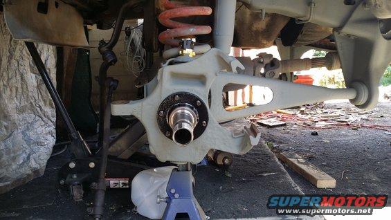 Close-up of vehicle's front suspension assembly, including the spindle, brake rotor area, coil spring, and control arms. Vehicle is on jack stands