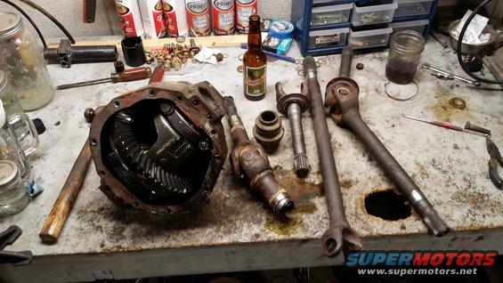 Differential housing and several axle shafts laid out on a workbench with tools and a beer bottle
