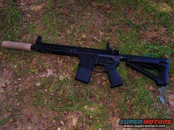 Black assault rifle with a tan suppressor lies on grass and dirt. The rifle has a black magazine and adjustable stock
