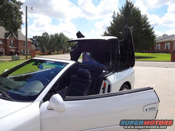 White convertible sports car, top mechanism partially open, driver's side door open, seen from the front passenger side