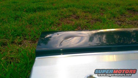 Dark green car tailgate with a large dent and chrome trim below