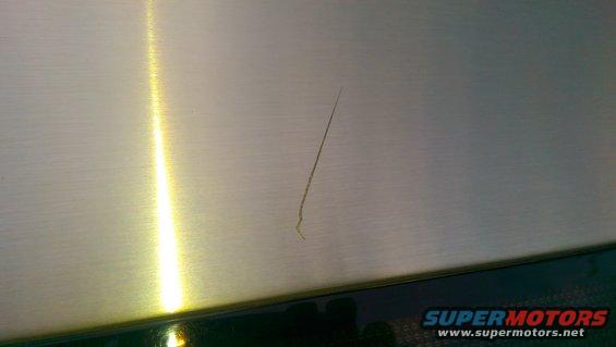 Close-up of a scratched metal surface with a bright light reflection and "SUPERMOTORS www.supermotors.net" text at the bottom