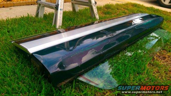 Dark green car tailgate trim piece with silver accent strip, lying on grass next to a ladder
