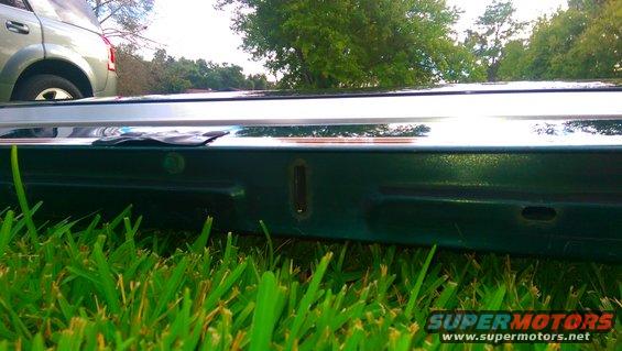 Close-up of a dark green vehicle tailgate with a chrome trim piece above it. Another vehicle is visible in the background
