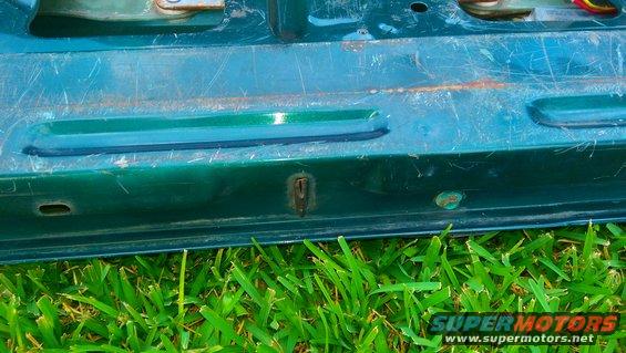 Green tailgate section with visible scratches and a rusty lock mechanism, grass in foreground