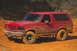 1995 Ford Bronco - A red Ford Bronco navigates a muddy terrain, its body caked in dirt.