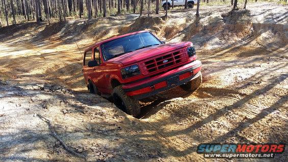 Red 1995 Ford Bronco navigating a steep dirt hill with one wheel off the ground. Another white SUV is visible in the background