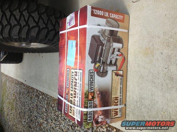 Box for a Badland 12000 lb capacity off-road winch with cable, next to a large tire