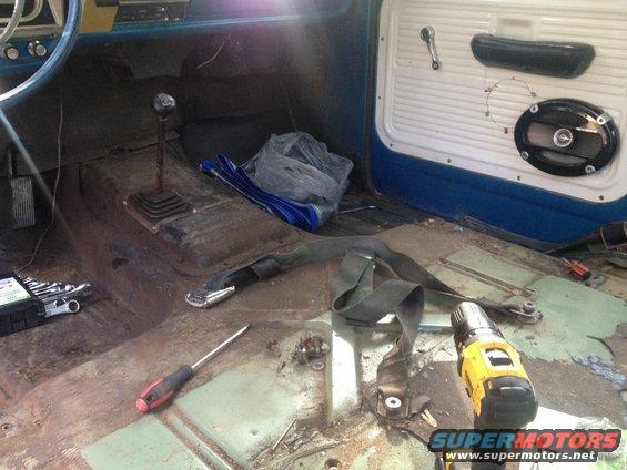Interior of a truck with floor removed, showing rust and tools. Seatbelt and gear shifter visible