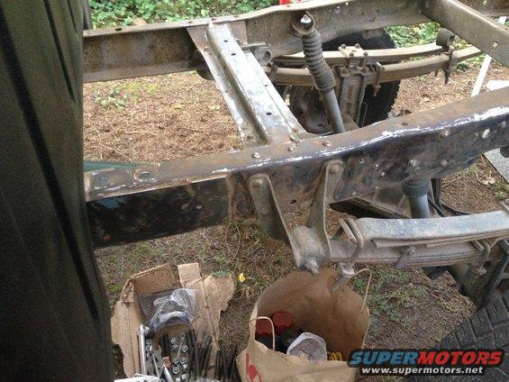 Close-up of a rusty truck frame with leaf springs and shocks. Parts are in boxes on the ground