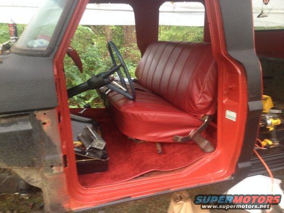 Interior view of a red truck cab showing a bench seat, steering wheel, and door panel. The truck appears to be undergoing restoration