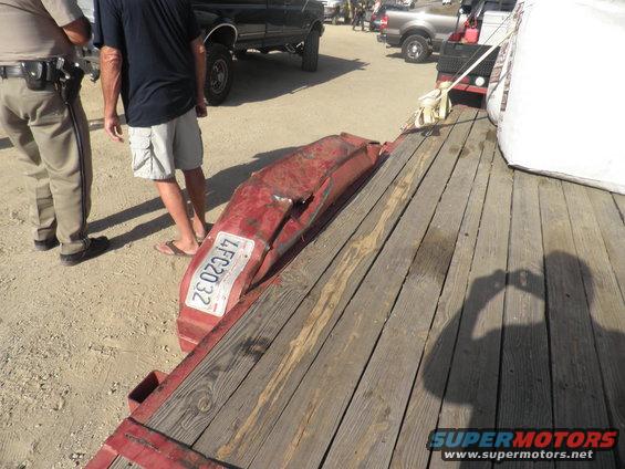 Damaged red bumper with license plate LFC2032 on a trailer, with people and trucks in the background