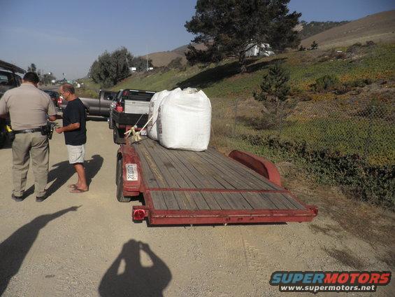 Red trailer loaded with two large white bags, parked on a dirt road next to a pickup truck