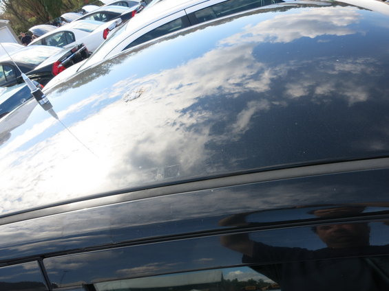 Reflection of clouds and a person on the dark roof of a car, with other cars visible in the background