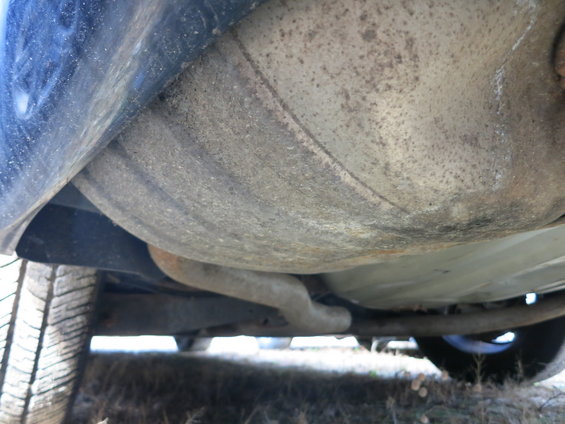 Underside of a blue vehicle showing the exhaust pipe and fuel tank, with a tire visible on the left